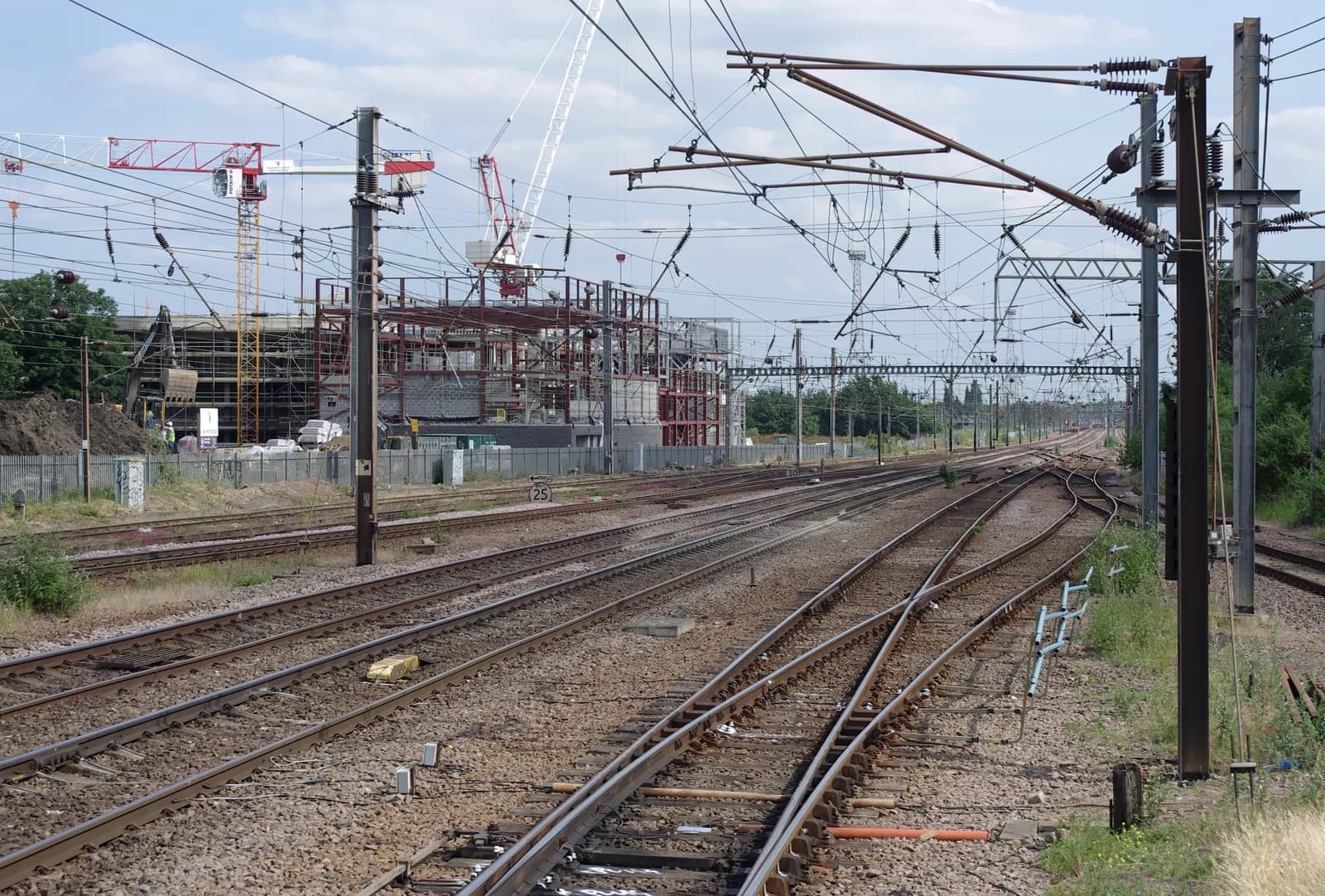 Multiple parallel train tracks with overhead power lines lead into the distance towards a construction site with cranes and buildings under development.